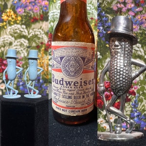 May include: A brown glass bottle of Budweiser Lager Beer with a white label. The label has a red and blue design with the Budweiser logo and the text "Does Not Contain Beer". There are two blue plastic peanut figures and a silver peanut figure with a top hat.