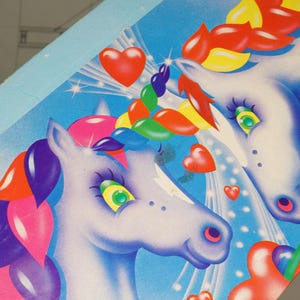 May include: A light blue box featuring a graphic illustration of two white unicorns with multicolored manes and tails. The design includes red heart shapes, starburst accents, and a bright blue background. The artwork displays a vibrant, colorful aesthetic.