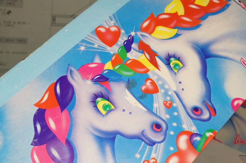 May include: A light blue box featuring a graphic illustration of two white unicorns with multicolored manes and tails. The design includes red heart shapes, starburst accents, and a bright blue background. The artwork displays a vibrant, colorful aesthetic.