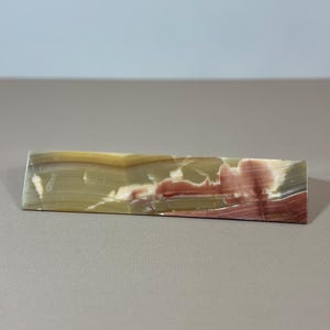 May include: A rectangular, polished stone with a layered appearance. The stone features bands of green, yellow, red, and white, creating a natural, marbled effect. The stone is approximately 6 inches long.