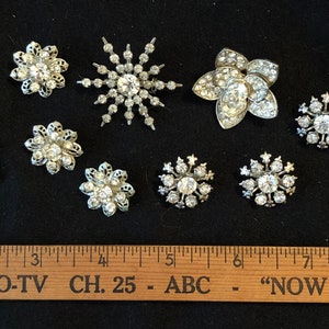 May include: Assortment of ten vintage rhinestone brooches in silver tone metal. The brooches feature various designs, including flowers, stars, and snowflakes. The brooches are arranged on a black surface with a wooden ruler.