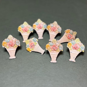 May include: A collection of eight vintage-style floral basket pins. Each pin features a cream-colored basket overflowing with colorful flowers in shades of pink, blue, yellow, and green. The pins are arranged on a dark green surface.
