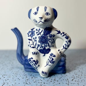May include: A white and blue ceramic teapot in the shape of a seated animal. The teapot features blue floral patterns and accents on the ears, spout, and base. The teapot has a whimsical design.