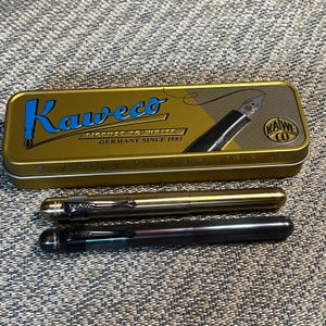 May include: A gold-colored rectangular metal tin with the blue text Kaweco, License to Write, Germany Since 1883, and a fountain pen illustration. Below the tin are two pens: one metallic gold with a black clip and one dark brown with a black clip.