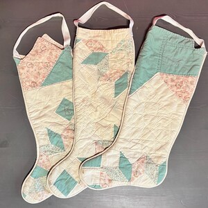 May include: Three vintage quilted Christmas stockings in white with pink and teal floral patterns. The stockings have a loop at the top for hanging.