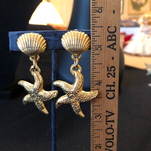 May include: Gold tone earrings featuring a seashell charm with a starfish dangling below. The earrings are approximately 2 inches long.
