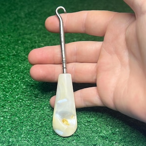 May include: A button hook featuring a metallic silver-toned shaft with a curved hook at the top and a smooth, iridescent white handle with subtle cream and gold-toned inclusions. The tool has a tapered, flat design for gripping and pulling buttons.