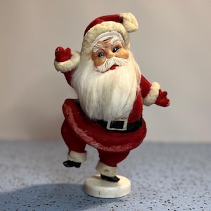 May include: A vintage-style Santa Claus figurine with a red velvet suit, white faux fur trim, and a black belt. The figure has a long white beard, blue eyes, and is posed standing on one leg on a round white base, with one arm raised in a wave.