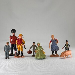 May include: A collection of miniature figurines depicting various figures. Included are a soldier, a hunter, a woman in a green dress, and a farmer. The figurines are painted in various colors and appear to be made of plastic.