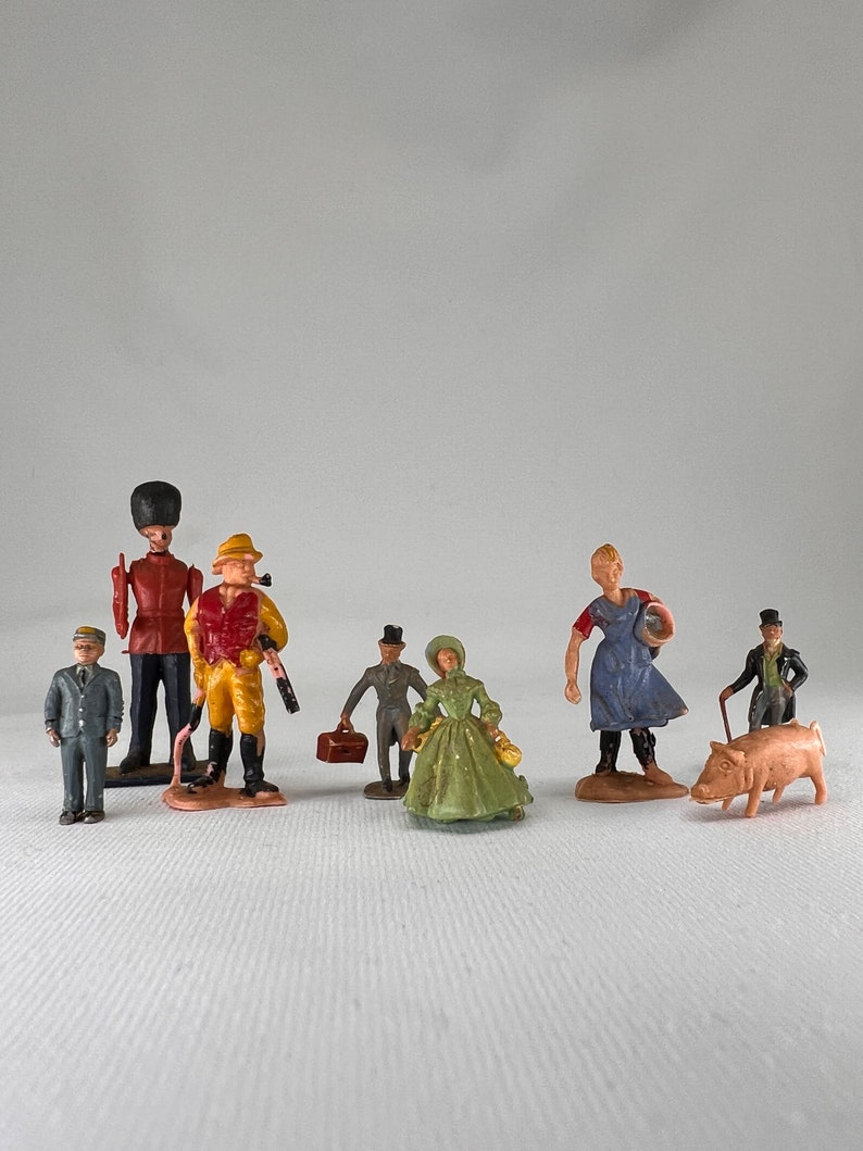 May include: A collection of miniature figurines depicting various figures. Included are a soldier, a hunter, a woman in a green dress, and a farmer. The figurines are painted in various colors and appear to be made of plastic.