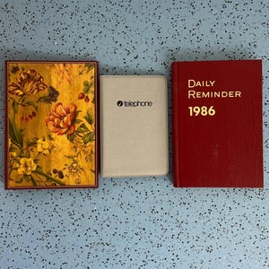 May include: Three books are displayed. A red book with "DAILY REMINDER 1986" in gold lettering. A gold and floral patterned book. A light gray book with the "telephone" logo.