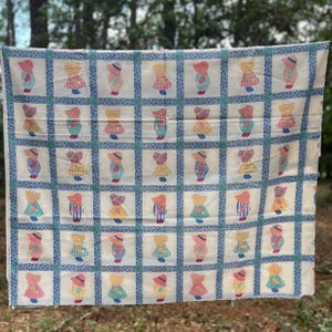 May include: A white fabric quilt featuring a repeating grid pattern of small, colorful figures wearing sun hats. The figures are arranged in rows and columns, separated by blue and white floral patterned borders that create a square block design.