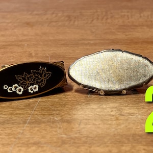 May include: Two vintage compact mirrors. The first mirror has a gold-colored metal frame with a black enamel background and a white floral design. The second mirror has a silver-colored metal frame with a textured surface.