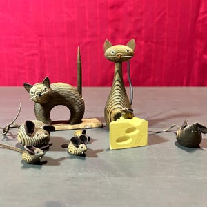 May include: A set of wooden cat and mouse figurines. The set includes two cats, one with a tail that stands upright and the other with a tail that lays flat. There are four mice, one of which is holding a piece of cheese. The figurines are all made of wood and have a natural wood finish.