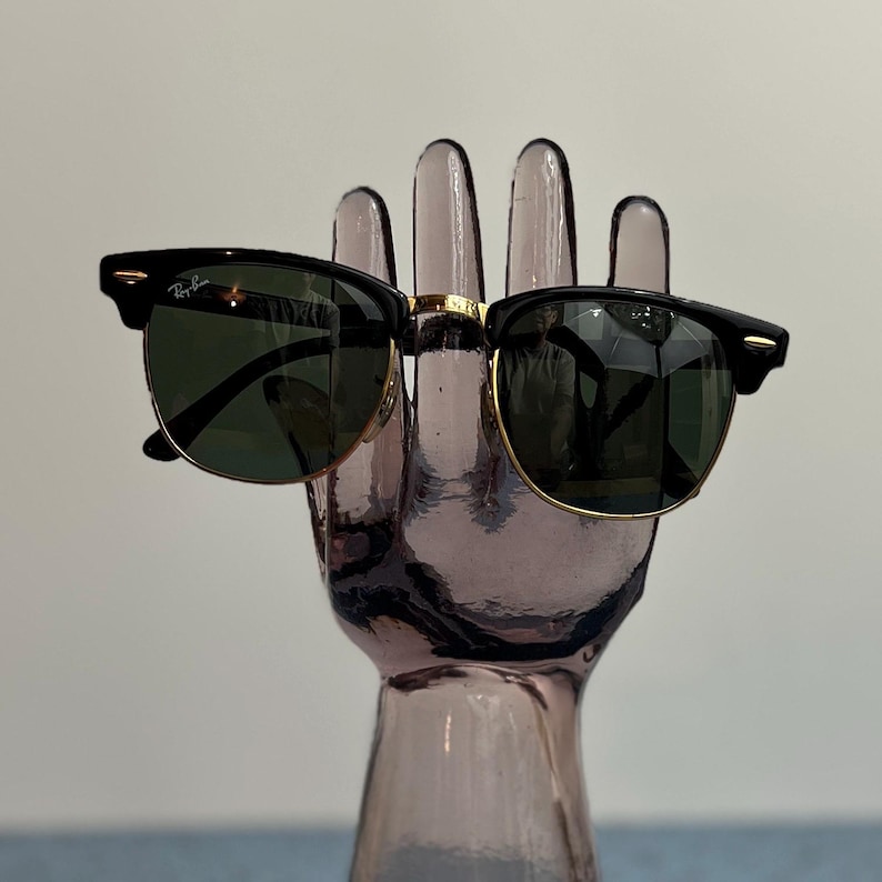 Op de afbeelding: Zwarte en goudkleurige zonnebril met donkergroene glazen. De zonnebril heeft een klassiek design met een zwart montuur en gouden accenten. Het Ray-Ban logo is zichtbaar op de linker lens. De zonnebril wordt getoond op een decoratieve hand.