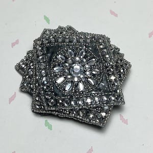 May include: A stack of square, silver-toned coasters. Each coaster features a central floral design with clear, faceted stones and is bordered by small, silver beads. The coasters have a dark gray fabric backing.