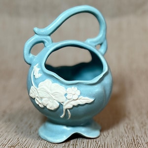 May include: A light blue ceramic basket with a handle and a round body. White floral relief designs adorn the front. The basket sits on a small, flared base. The handle curves upward and then back down.