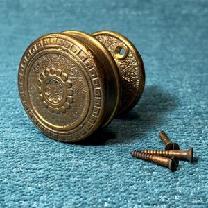 May include: Antique brass door knobs with intricate floral and geometric patterns. The knobs are round with a detailed design on the face and a circular backplate. Three screws are included. The hardware is likely for a vintage door.