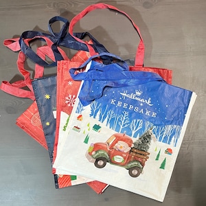 May include: Four reusable shopping bags with a Christmas theme. The bags are red, blue, and white with snowflakes, a snowman, and a truck with a Christmas tree. The bags are made of a lightweight material and have handles.