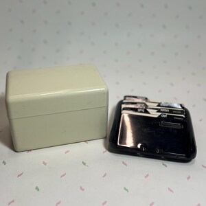 May include: A vintage cream-colored rectangular box with a hinged lid next to a black address card file. The address card file has labeled tabs and a glossy finish. The background is a light pink and white patterned surface.