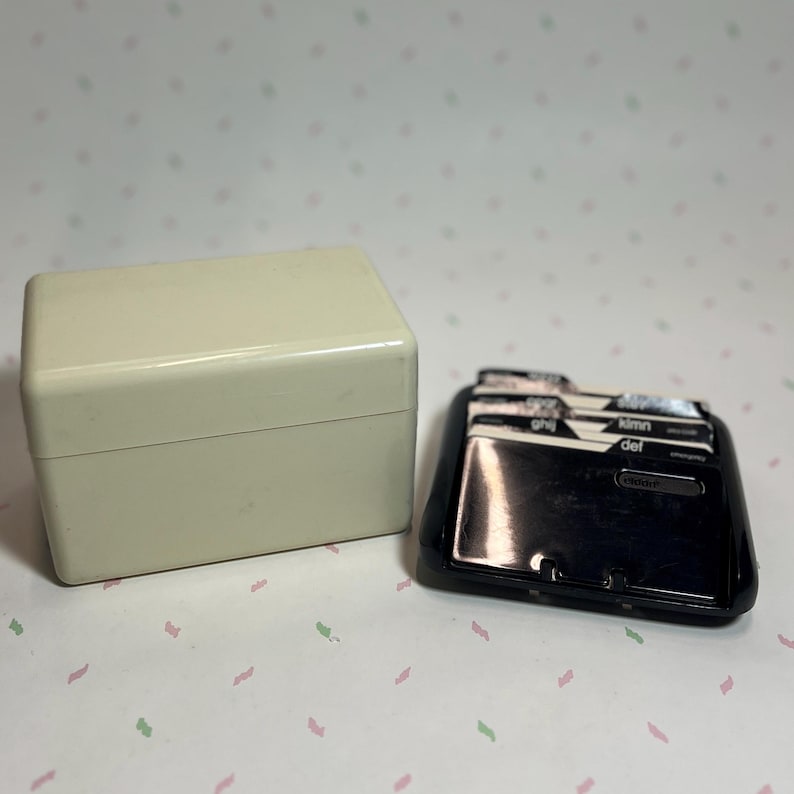 May include: A vintage cream-colored rectangular box with a hinged lid next to a black address card file. The address card file has labeled tabs and a glossy finish. The background is a light pink and white patterned surface.