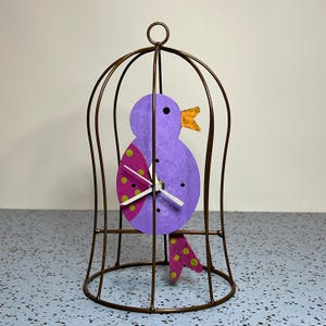 May include: A decorative clock shaped like a purple bird with a pink wing featuring yellow polka dots, an orange beak, and white hands, enclosed within a bronze-colored wire birdcage frame with a circular top loop for hanging or display.