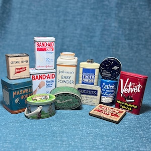 May include: A collection of vintage tins and containers, including Band-Aid boxes, Johnson's Baby Powder, and a Maxwell House coffee tin. Other items include a Velvet tobacco tin and a Carter's Midnight tin. The items are arranged on a blue surface.