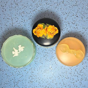May include: Three decorative vintage containers. One is a black oval with yellow roses, another is a light green round container with white doves, and the third is a peach-colored round container with a yellow spoon-shaped object.