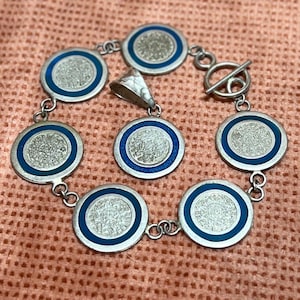 May include: A silver bracelet with a blue enamel design featuring the Aztec calendar. The bracelet has a toggle clasp and a pendant.