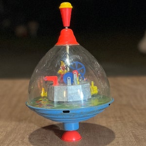 May include: A vintage toy top with a clear plastic dome, a red and yellow top, and a blue base. Inside the dome, a small figure is operating a miniature machine.