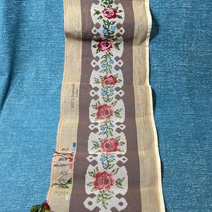 May include: A needlepoint canvas featuring a floral design with red roses, blue accents, and green leaves. The canvas has a brown central panel with a cream-colored border. Colorful embroidery floss is visible at the bottom.