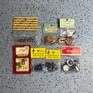 May include: A collection of eight miniature accessory sets in clear plastic bags with cardboard headers. Items include gold-toned metal kitchenware, blue and white ceramic-style dishes, and small grocery items labeled Fibre-Craft and Concord.
