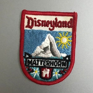 Puede incluir: Un parche bordado rojo, blanco y azul con el texto "Disneyland" y "Matterhorn" en negro. El parche presenta una monta&ntilde;a blanca con un cielo azul y un sol amarillo.