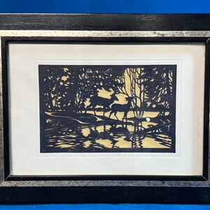 May include: Framed paper cut art depicting deer in a forest scene. The artwork features a black silhouette of deer, trees, and water against a light background. The frame is black with a silver trim.