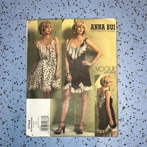 May include: An Anna Sui Vogue Patterns sewing pattern envelope labeled V1104. The front displays three images of a dress with ruffled straps and hemline, shown in both a patterned fabric and a solid black fabric, with the text Anna Sui Vogue Patterns.