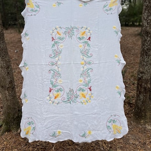 May include: White tablecloth with embroidered floral design. The embroidery features yellow, green, and red accents. The scalloped edges add a decorative touch. The tablecloth is hanging between two trees.