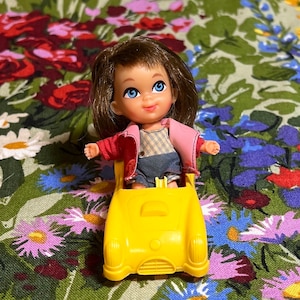 May include: A small doll with brown hair and blue eyes, wearing a red jacket, a plaid shirt, and denim shorts, sits in a yellow toy car. The doll is smiling. The background is a floral pattern.