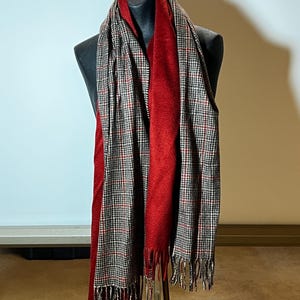 May include: A long scarf featuring a reversible design with one side in a solid deep red color and the other side in a black, white, and red houndstooth pattern. The scarf has a soft texture and is finished with fringe detailing at both ends.