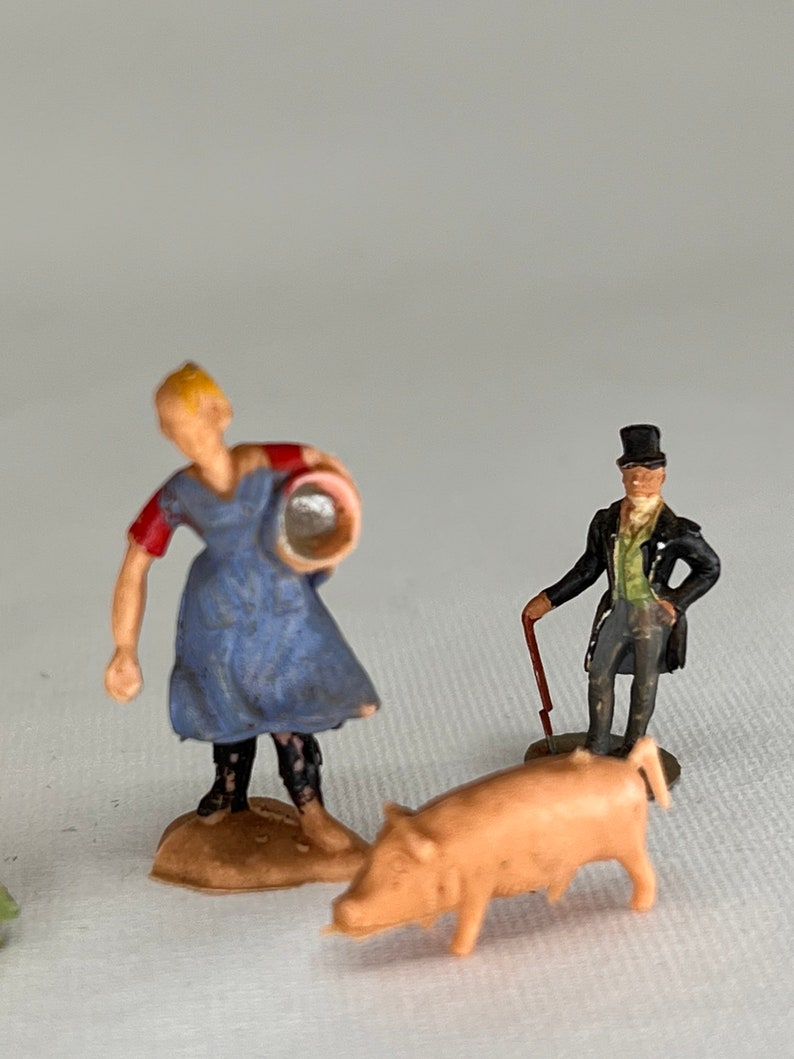 May include: Miniature figurines of a farmer, a gentleman, and a pig. The farmer wears a blue dress and red sleeves, holding a bucket. The gentleman is in a black coat and top hat, with a cane. The pig is light pink.