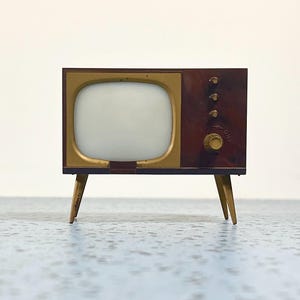 May include: A miniature television set with a dark brown cabinet, a gold-toned screen frame, and three small knobs above a larger dial labeled ON. The device is supported by four thin, angled gold-colored legs and features a white screen.