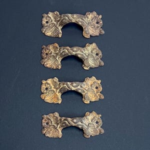 May include: Four antique bronze drawer pulls with a floral and leaf design. Each pull has a curved handle and two mounting holes. The pulls are arranged vertically against a dark background. The bronze finish has a vintage, aged appearance.