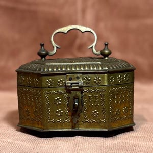 May include: A small, octagonal, brass box with an ornate design. The box has a hinged lid with a decorative latch and a handle on top. The box is decorated with floral patterns and has a slightly aged appearance.