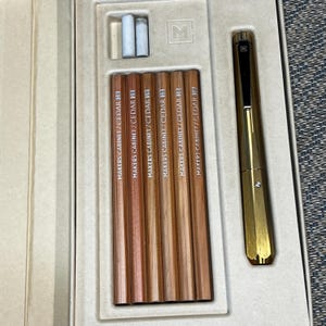 May include: A set of six wooden pencils labeled Makers Cabinet Cedar 2B, two small white erasers, and a gold-toned metal mechanical pencil with a black clip, all housed in a fitted beige tray with an embossed M logo on the interior surface.