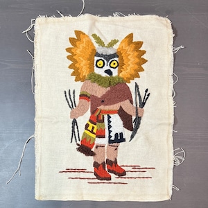 May include: A beige fabric wall hanging with an embroidered owl wearing a brown and white feathered headdress. The owl is holding a bow and arrow in each hand. The background is a beige fabric with a brown border.
