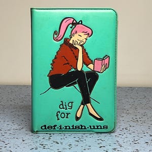 May include: A teal rectangular notebook featuring a graphic illustration of a person with pink hair wearing a rust-colored sweater and black pants, reading a pink book. The text "dig for def-i-nish-uns" is printed in black at the bottom.