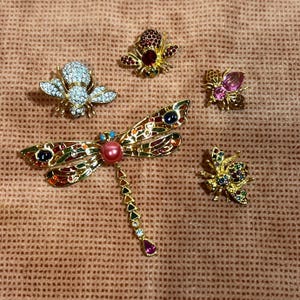 May include: Five gold-tone brooches with colorful rhinestones. The brooches feature a dragonfly, a bee, a butterfly, and two other insect designs.