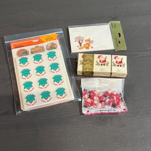 May include: Assortment of Christmas and Thanksgiving themed craft supplies including stickers, party matches, and small Santa figurines. The stickers feature a green wreath with the text "You're Tops!" and a turkey with the text "Happy Holidays". The party matches feature a Santa Claus illustration and the text "Party Matches". The Santa figurines are red and white with black facial features.