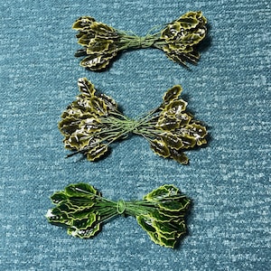 May include: Three bundles of artificial green holly leaves tied with green wire. The leaves have a glossy finish and are arranged in a bow-like shape. The background is a textured teal fabric.