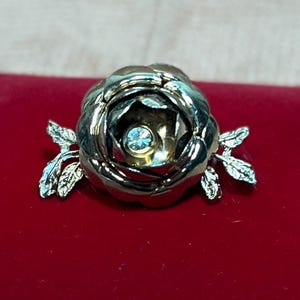 May include: A gold-toned rose brooch with a clear rhinestone in the center. The rose has a textured, layered design and is accented with two small leaves on either side.