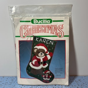 May include: A Bucilla Christmas needlepoint stocking kit. The kit features a green stocking with a teddy bear dressed as Santa Claus. The name "KAREN" is stitched on the stocking. The kit is in a clear plastic package.